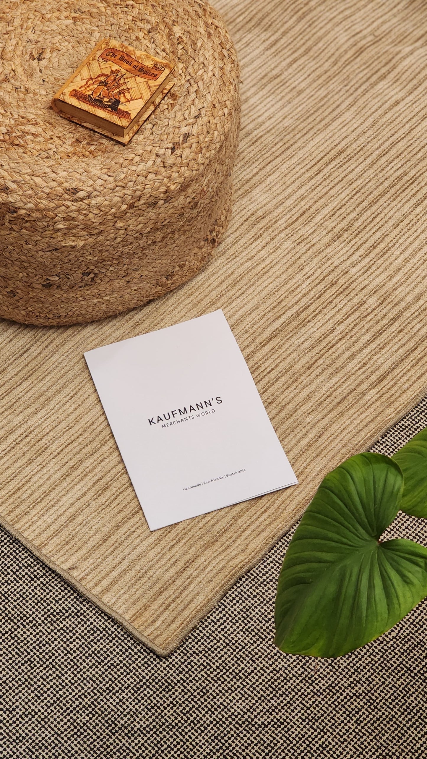 A beige cotton rug with a flatweave pattern, placed on a floor next to a round jute rug, a cardboard box with a design, and a business card with the text 'Kaufmann's'. The background is out of focus.